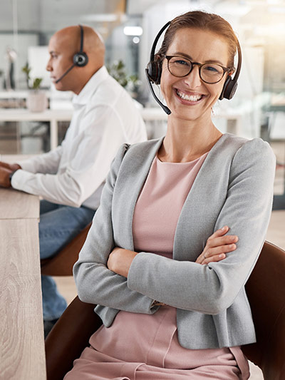 Woman at desk wearing a headset and microphone. She is wearing a dress and jacket and smiling at the camera with her arms crossed.